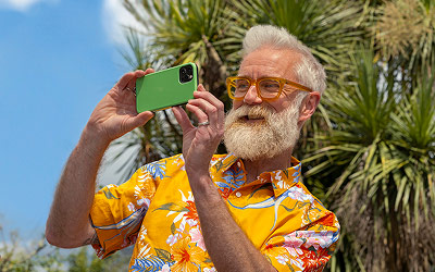 A person in a bright floral shirt holding up a green phone in front of palm trees.
