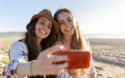 A person holding a red smartphone while taking a selfie with another person on a sandy beach.